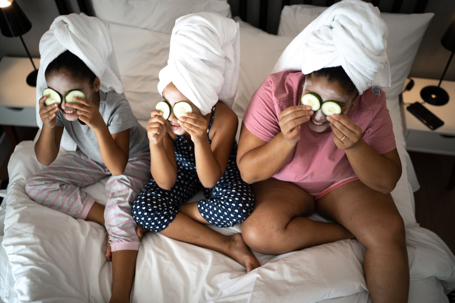 Två flickor och en kvinna sitter i en säng med handukar virade runt håret och gurkor för ögonen i ett hemmaspa