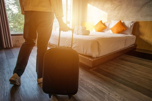 Hotellrum i solljus. En person går mot sängen med en rullresväska.